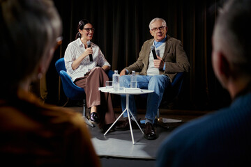 Wide shot of senior Caucasian man and young woman speaking into microphones during panel...