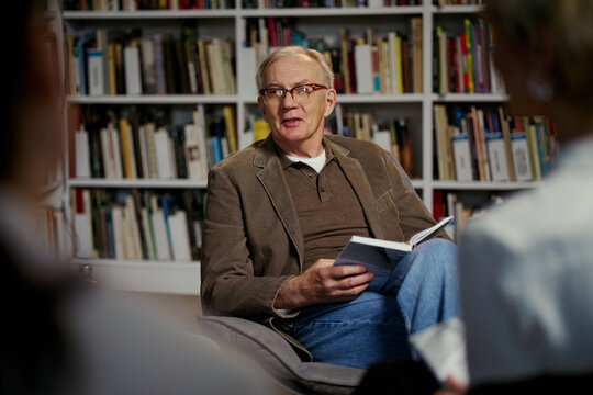 Senior Caucasian man reading from book and speaking to group in library, leading discussion and sharing knowledge. Useful for education, mentoring, book club, workshop marketing