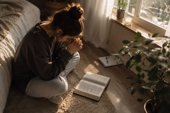 Young Christian woman praying beside an open Bible while sitting on the bedroom floor in warm morning sunlight, personal faith and devotion concept.