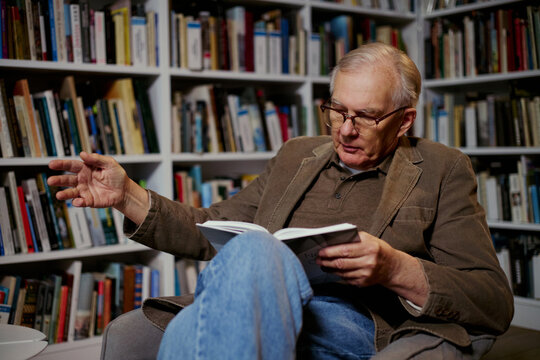 Senior Caucasian man reading book and gesturing while explaining ideas in home library, conveying expertise and lifelong learning. Useful for education, publishing, mentoring themes