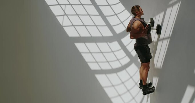 Athletic man lifting weights, in a bright, shadow patterned studio, focusing intensely on his workout. Athletic Man Doing Strength Training in Sunlit Studio. Vertical video.