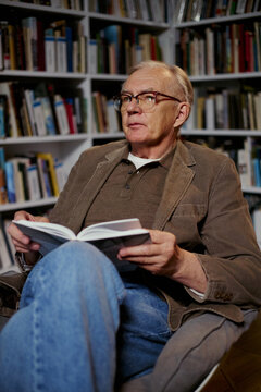 Vertical shot of senior Caucasian man reading book while sitting in home library, looking away and reflecting on knowledge, learning, retirement lifestyle. Useful for education or publishing