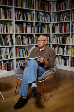 Vertical shot of senior Caucasian man reading book while sitting before bookshelves, suggesting lifelong learning and knowledge. Useful for education, publishing, retirement content