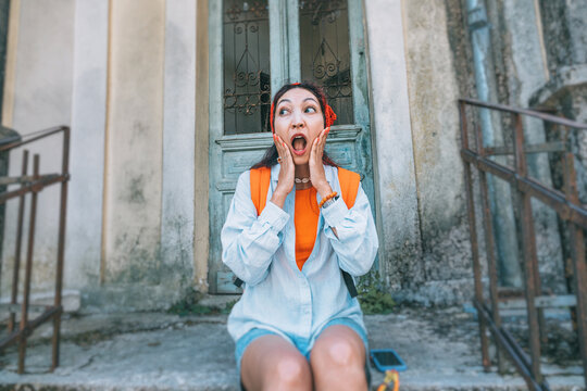 Shocked young woman traveler with hands on face sitting on steps of an abandoned building