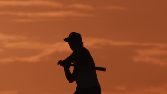 A silhouette of a man against an orange sunset holds a baseball bat, throws it up, catches it and strikes it