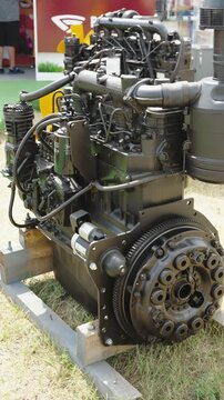 A brand new black diesel engine with an attached clutch assembly stands on a wooden frame at an outdoor exhibition.