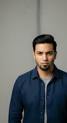 Obraz premium Young Middle Eastern man with dark hair and beard wearing navy button-up shirt over gray undershirt, posed against neutral gray wall for portrait and headshot use.