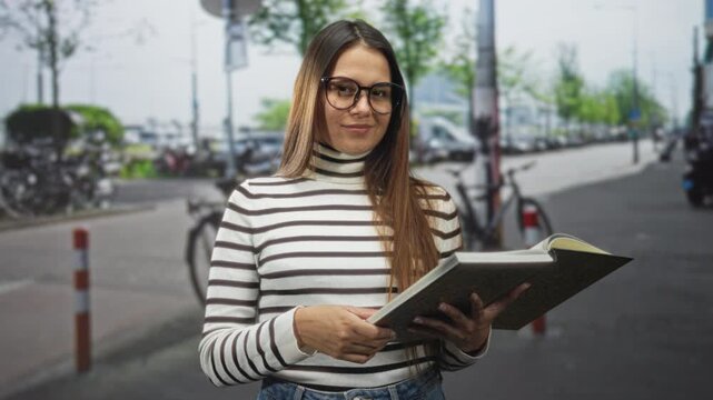 Woman holding open book and pointing finger with visible hand on street near bicycles and parked cars, wearing striped turtleneck and glasses; curiosity learning focus.