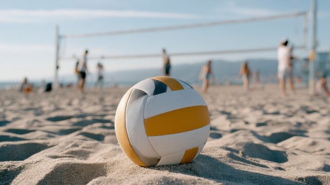 A volleyball rests on sandy beach with players and a net blurred in the background under a clear sky.