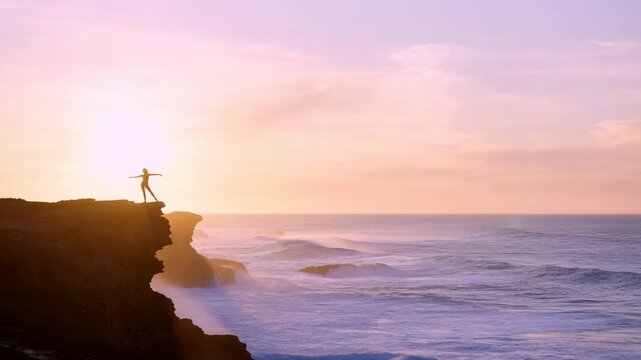 Silhouette on rugged coastal cliff at sunrise with outstretched arms facing pastel sky, glowing ocean horizon, soft clouds, dramatic spray, and peaceful scenic balance