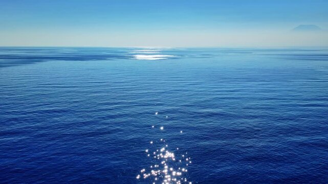 Panning shot captures a sparkling sunlit path across a peaceful deep blue ocean surface.