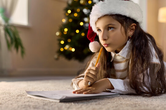 Christmas child writing letter to Santa at home. Girl lies on floor by festive tree in santa hat, focused on wishlist with pen. Winter holiday and New Year celebration and preparations concept.