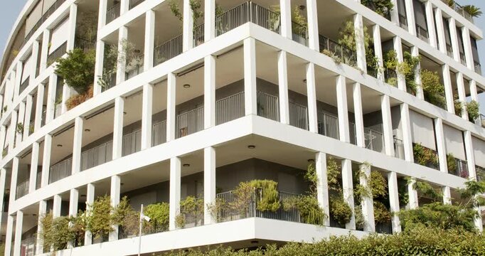 A modern residential building featuring white structural columns, open balconies decorated with green potted plants and vegetation, creating a eco friendly urban architectural style on a bright day.