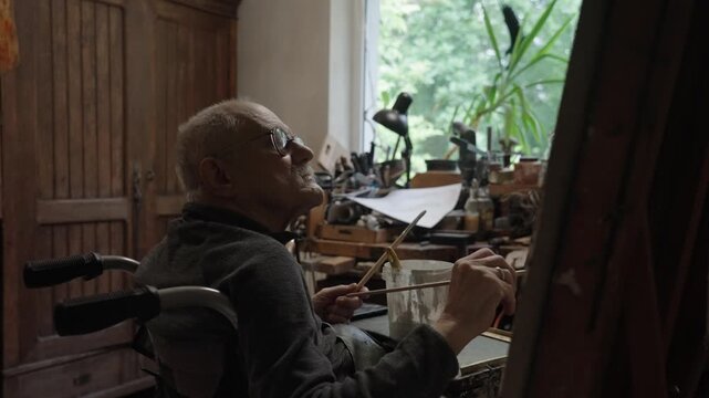 Senior artist on a wheelchair paints an oil painting on canvas in a studio