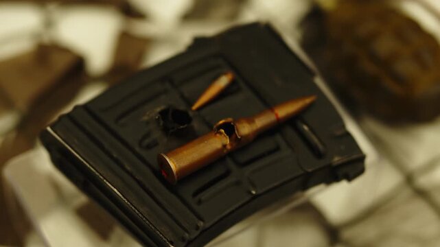 A rifle cartridge with a shrapnel or bullet hole through its casing rests on a damaged black rifle magazine.
