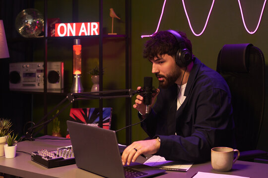Young man hosting live podcast, speaking into microphone while monitoring laptop and audio mixer in studio. Ideal for streaming, broadcasting, creator economy, media production campaigns