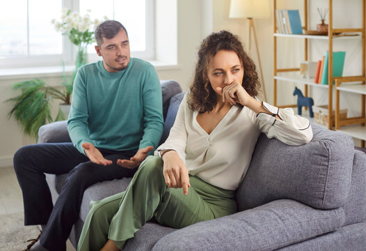 Husband talking with his unhappy upset wife sitting on sofa at home. Young married couple quarreling and arguing in the living room. Marriage problems, relationship and divorce concept.