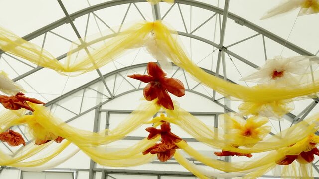 Yellow and orange fabric flower decorations hang from the metal trusses of a large event tent ceiling.