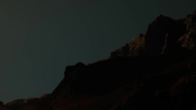 A mountain stands in the dark at night under a sky with minimal light. The outline of the peak is visible against the dark background. The scene captures the stillness of the night.