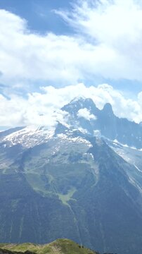 Hiker woman with backpack standing on a cliff edge enjoying the stunning view of a mountain valley, with camera revealing the alpine landscape below and snow-capped peaks