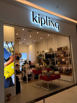 A retail store showcasing Kipling bags and accessories. The store features bright lighting and a modern design. Shelves display a variety of colorful handbags and backpacks.