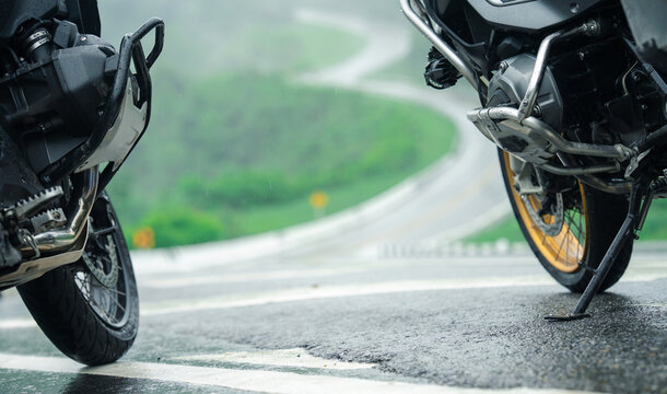 Two motorcycles parked on wet, winding road with lush green scenery in background, evoking sense of adventure