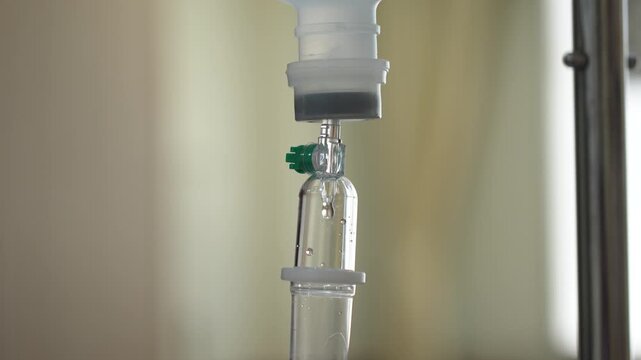 A close-up shot of an intravenous (IV) drip infusion set with saline solution drops in a hospital room, featuring a blurred background.	