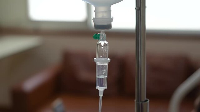 A close-up shot of an intravenous (IV) drip infusion set with saline solution drops in a hospital room, featuring a blurred background.	