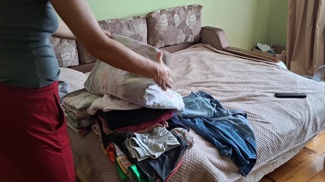 Woman folds clothes on a bed in a room with light colors and a sofa