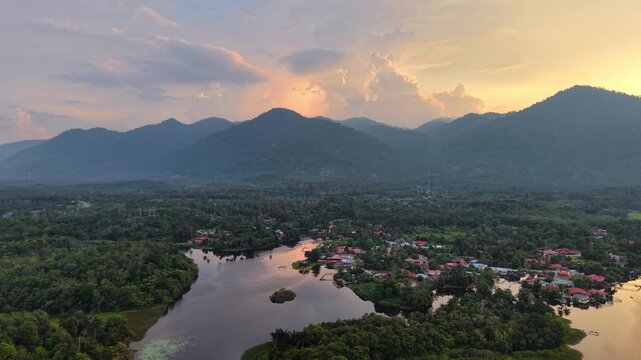 A peaceful aerial view of a village nestled among green forest along a river at sunset with mountains in the background and calm water reflecting the sky at Tasik Raban