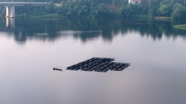A calm lake features a small wooden boat next to aquaculture cages surrounded by dense green forest and clear water at Tasik Raban