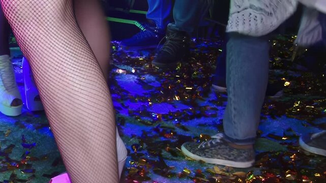 Lively partygoers move in rhythm while wearing stylish fishnet tights and high heels on a confetti covered floor in a dimly lit club at night surrounded by neon laser beams and energetic music