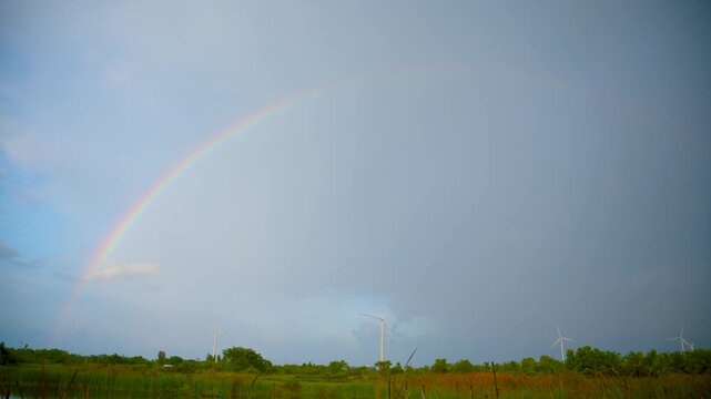 Beautiful Full Rainbow Arching Over Green Fields with Wind Turbines Against Gray Blue Sky After Rain, Renewable Energy Landscape Thailand