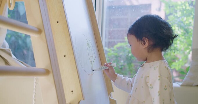A cute toddler girl proudly smiles at the camera while drawing on a whiteboard easel, engaging in creative play for her early childhood development and homeschooling.
