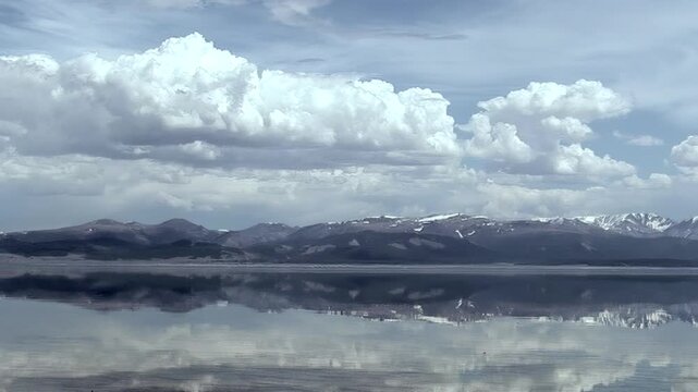 Khuvsgul Lake in Mongolia presents a spectacular reflection. Towering mountains, some with snow, and a sky filled with dramatic white clouds are perfectly mirrored on the tranquil water.
