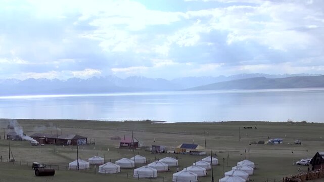 Charming nomadic gers stand pristine Lake Khuvsgul in Mongolia. Smoke drifts from dwellings, showing daily life and warmth. Vast, majestic landscape with distant mountains unfolds under cloudy skies.