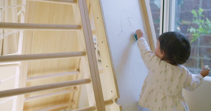 A young toddler erases her shark drawing from a whiteboard easel in a sunlit playroom, developing fine motor skills and exploring her creativity through independent play.