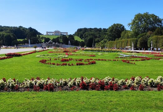 Vibrant summer flower arrangements and green lawns in Schonbrunn Palace gardens. The iconic Gloriette pavilion stands on a hill in the background under a blue sky.