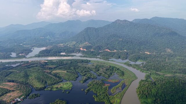 Aerial shot capturing a green river bend surrounded by dense forest and a mountain range under a partly cloudy sky during daylight at Tasik Raban