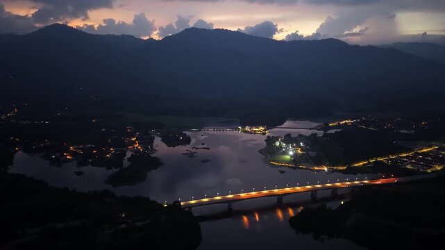 Dusk paints mountains in soft purple hues. River reflects city lights and distant village glow at Tasik Raban