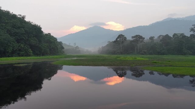 Calm lake reflects distant mountains and soft clouds. Mist hangs low over valley at Tasik Raban