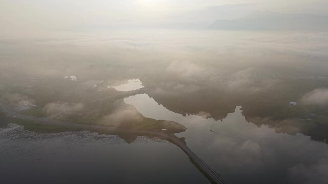 Misty aerial view of a coastal landscape featuring a long bridge crossing calm water with fog at Tasik Raban