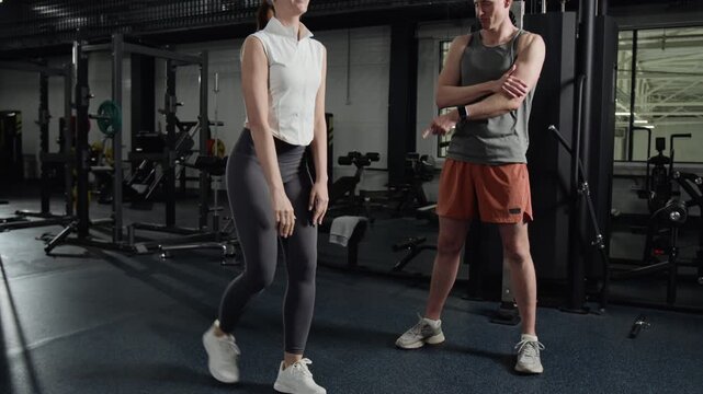 Tilt up shot of young woman performing lunges in gym while male personal trainer guides her. Clip for fitness center promotions and personal training advertisements