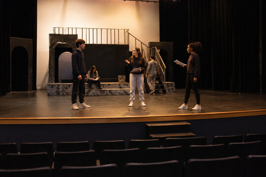 High school students rehearse a dramatic scene with scripts on a theater stage under warm lights while empty auditorium seats fill the foreground.