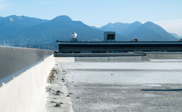 Flat roof parapet with granule loss on modified bitumen cap sheet. Aggregate pile visible at base. SBS 2-ply roofing system requiring maintenance. North Shore Mountains background. Selective focus.