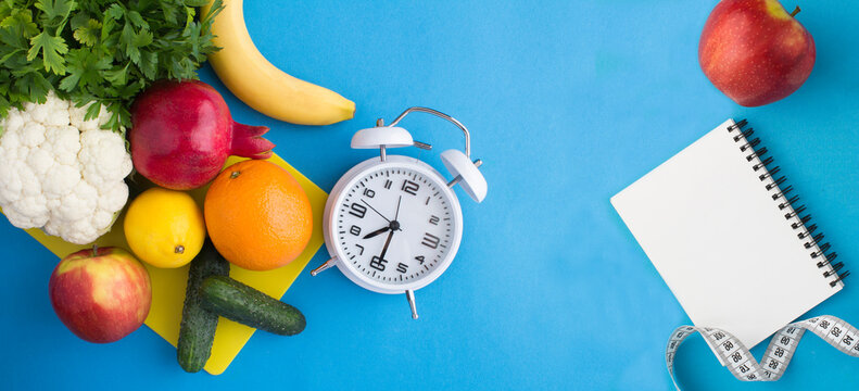 White alarm clock, vegetables, fruits and notepad for text on a blue background. Top view.