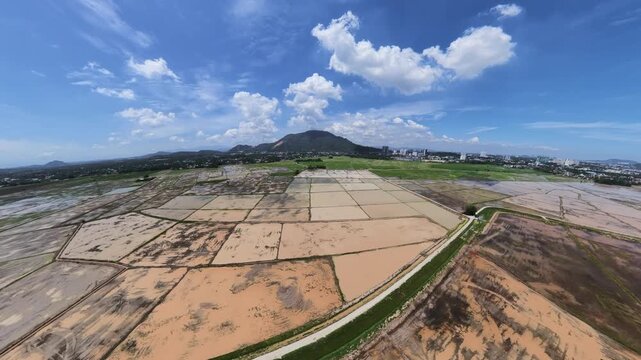 A wide aerial view of farmland divided into plots with a distant city skyline, surrounding hills, and a partly cloudy sky under bright sunlight.