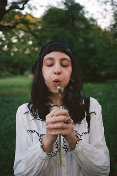 Woman blowing on dandelion in a green field during sunset near trees