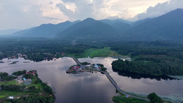 A tranquil aerial view of a lake surrounded by dense forest and mountains during sunset, featuring a narrow bridge connecting the land at Tasik Raban