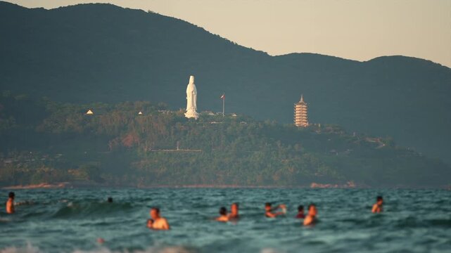 People swim in Danang sea, Lady Buddha statue and Linh Ung Pagoda above waves and tourists in Da Nang, Coastal view of Son Tra Peninsula with Buddhist landmark, mountains and ocean shoreline 4k